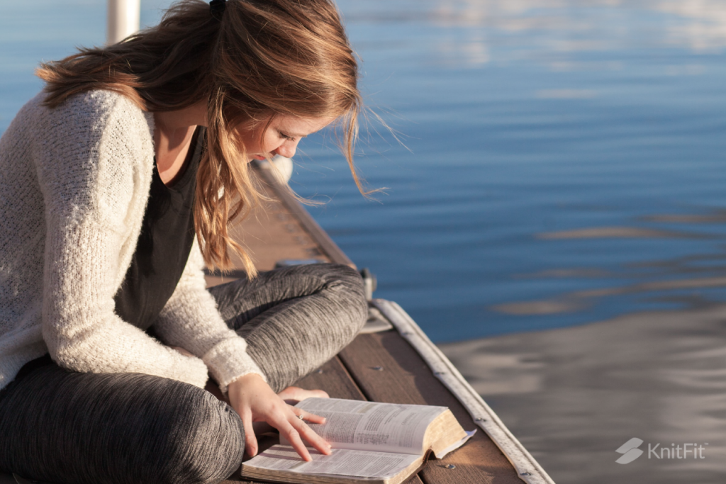 A woman reads a book to learn skills for knitting the unshaped sweater or garment, sitting on a dock by the sea