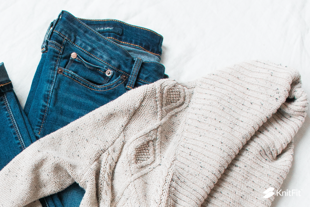 A cream-colored, boxy, loose sweater with cable designs lies over a pair of folded blue jeans. 